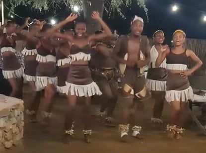 A group of dancers in traditional attire performing energetically outdoors at night. The dancers wear brown and white outfits adorned with fringe. String lights hang above, and a tree is visible in the background, adding to the festive atmosphere.