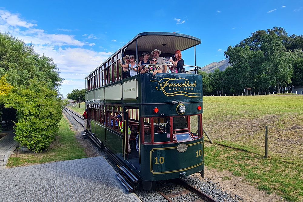 A vintage double-decker tram filled with passengers travels through a scenic landscape. The tram, labeled 'Franschhoek Wine Tram,' has an open top where tourists enjoy the ride under a clear blue sky. Lush greenery and a mountain backdrop complete the picturesque view.