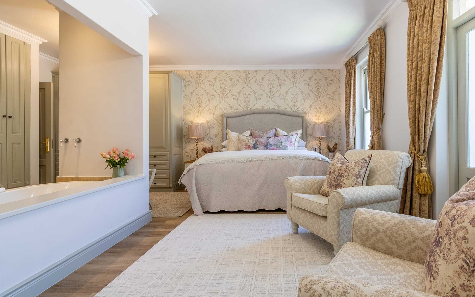 A serene bedroom with a super king bed adorned with floral pillows, soft lighting, and elegant wallpaper. Cozy armchairs and light-filled windows enhance the inviting atmosphere. A bathtub and fresh flowers add to the luxurious, restful appeal of this private villa retreat.
