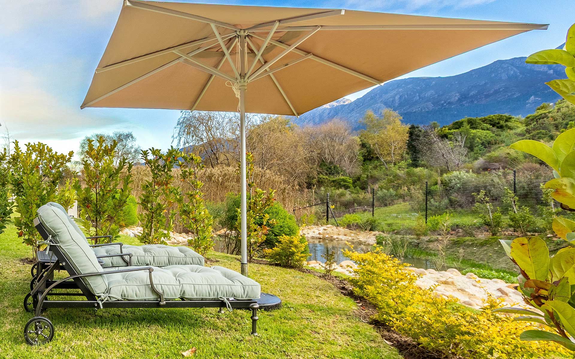 Two lounge chairs under a large umbrella sit on a grassy lawn beside lush gardens. A peaceful mountain stream flows nearby, with mountain views in the background, creating a serene outdoor setting at Villa 56.