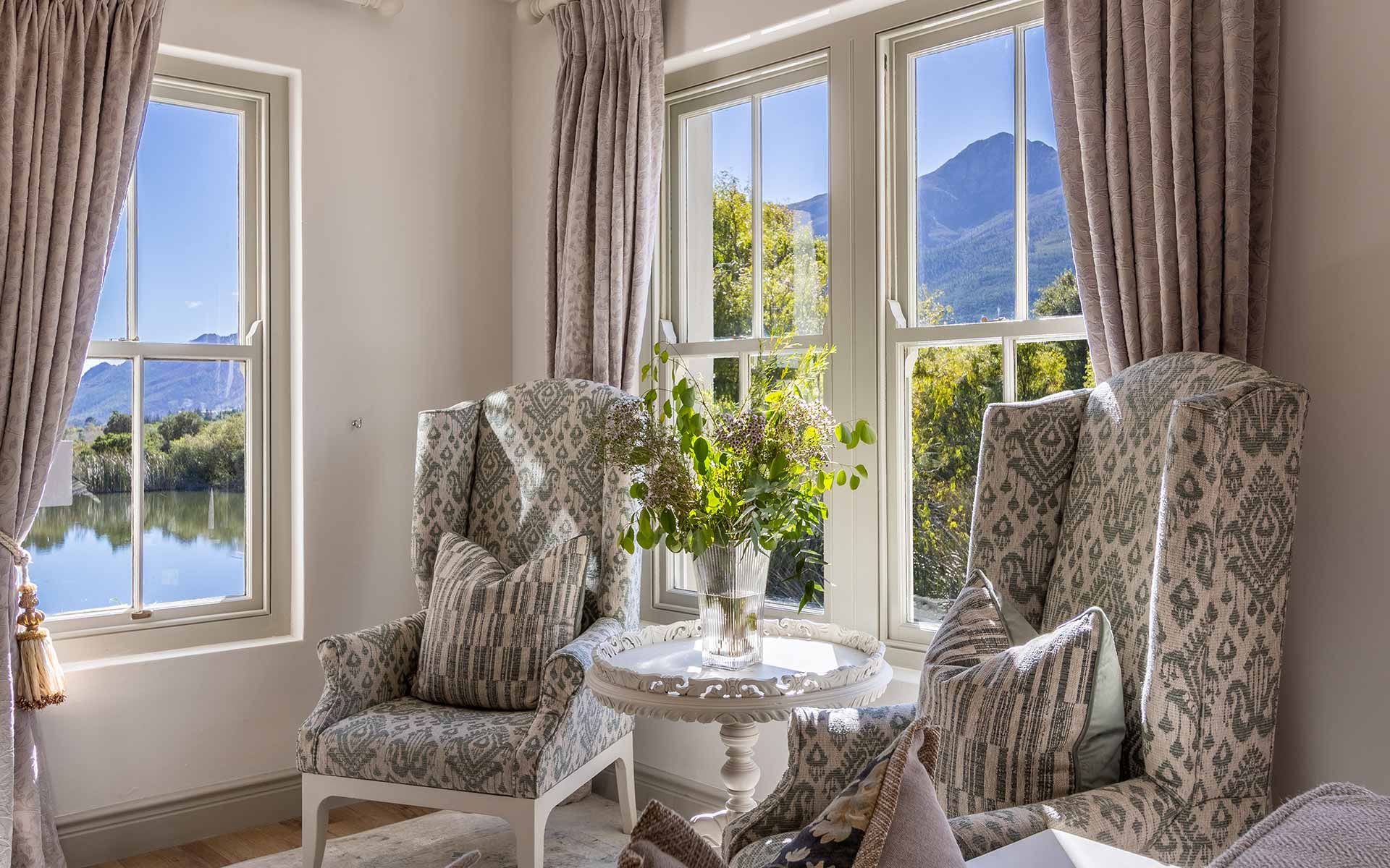 A cozy sitting alcove in Villa 31 with two patterned armchairs, a round table with a vase of flowers, and large windows revealing stunning lake and mountain views. Soft natural light enhances the serene and elegant atmosphere, perfect for relaxation.