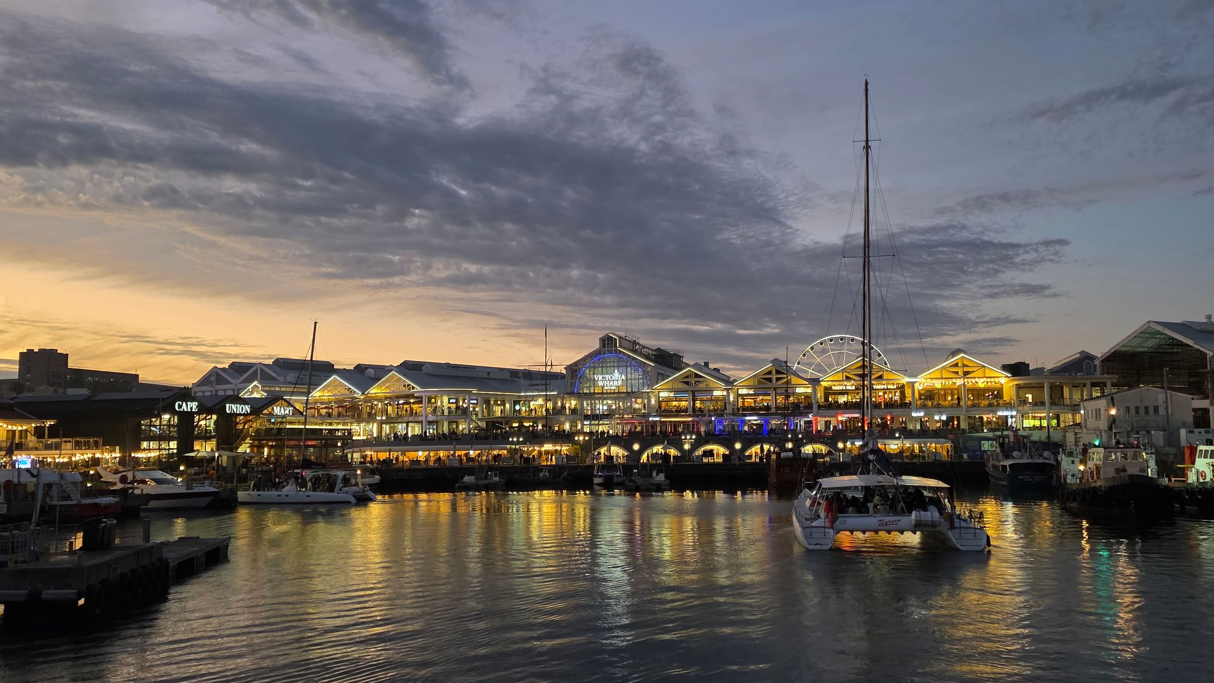 The V&A Waterfront in Cape Town at twilight, showcasing illuminated shops and restaurants along the harbor. Boats are docked in the foreground, while a colorful sky and scattered clouds create a picturesque backdrop, capturing the vibrant yet serene atmosphere of this popular destination.