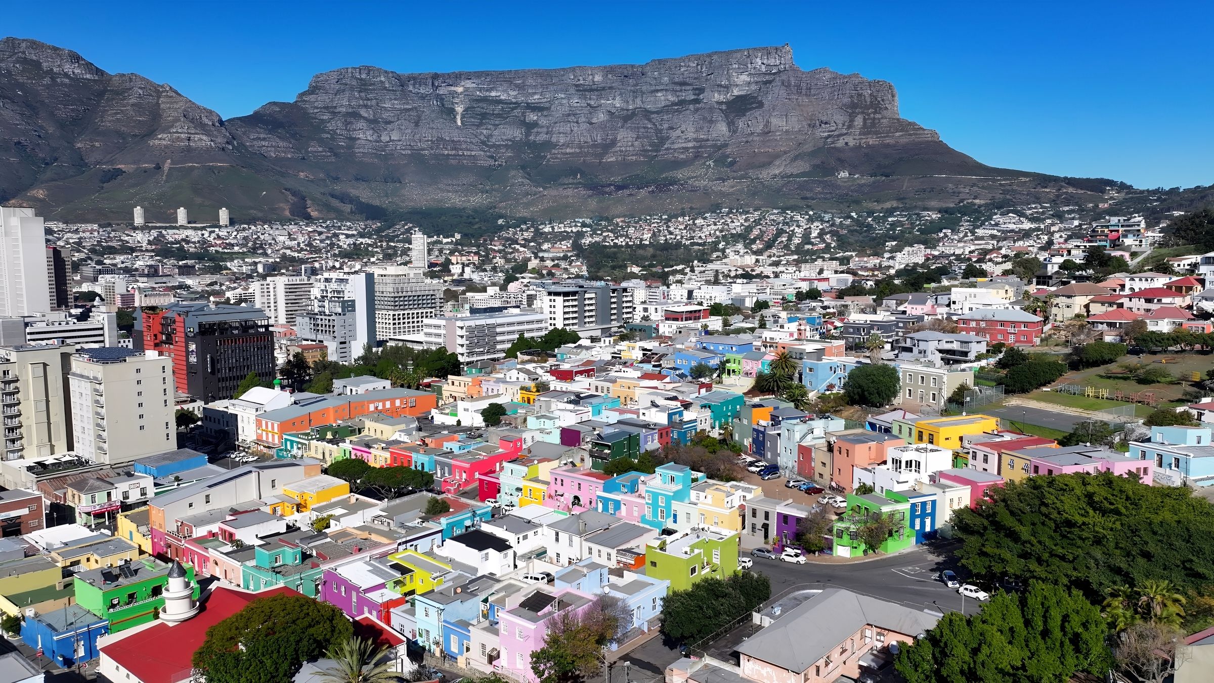 A panoramic view of Bo-Kaap, Cape Town, featuring a vibrant array of brightly painted houses in colors like pink, green, and blue. The historic Cape Malay quarter is set against the backdrop of Table Mountain, showcasing cobblestone streets and the rich cultural heritage of the community.