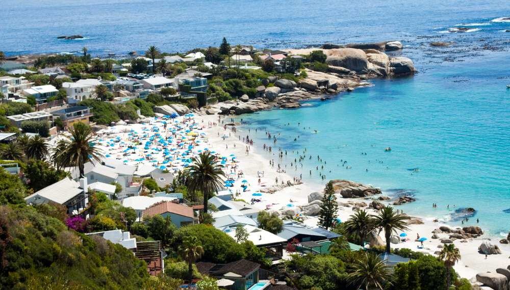 Aerial view of Clifton Beach in Cape Town, showcasing the vibrant beach culture with colorful umbrellas and sunbathers. The scene includes turquoise Atlantic waters, white sandy shores, lush greenery, and the rocky coastline, capturing the dramatic natural beauty against the urban backdrop.