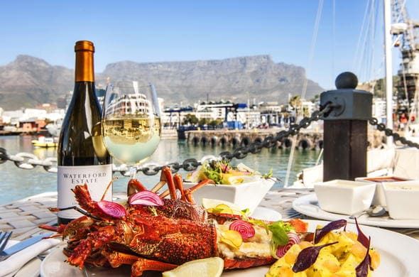 A scenic view of a seafood meal featuring lobster, a bottle of white wine, and a glass, set on a table by a harbor. The background showcases a mountain range and a marina with boats under a clear blue sky.