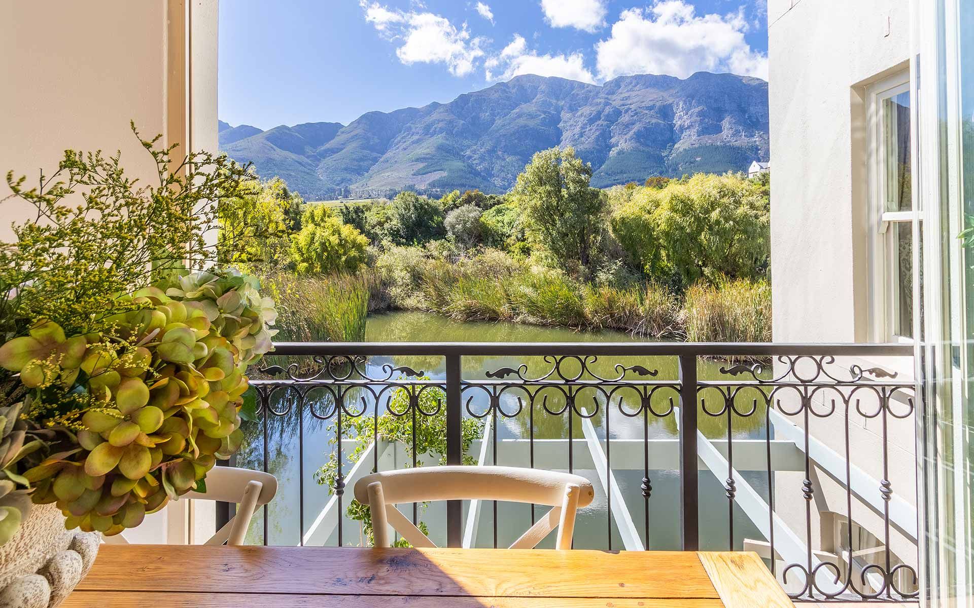 View from a villa balcony overlooking a serene lake, surrounded by lush greenery and framed by majestic mountains under a clear blue sky. A wooden table with a potted plant is visible in the foreground, enhancing the tranquil and picturesque setting.