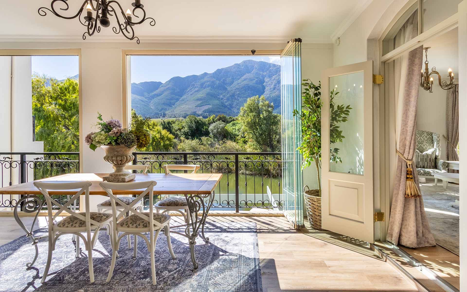 Elegant dining area in Villa 31 with a wooden table and chairs, overlooking Patrysfontein Lake. Large windows frame the Mont Rochelle mountains, offering a stunning natural backdrop. A potted plant and floral arrangement add charm to the serene, sunlit space.