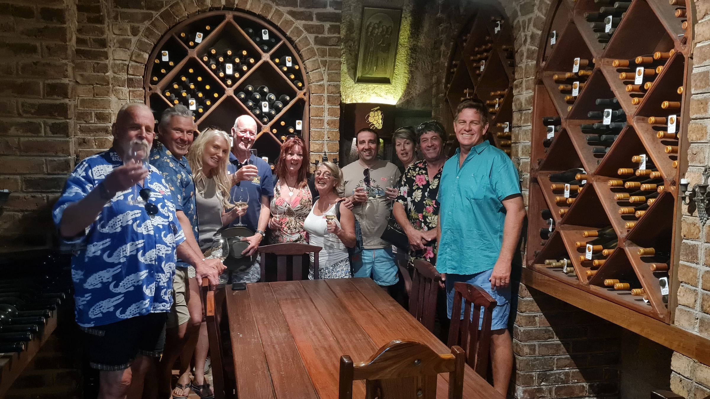A group of people standing in a wine cellar, holding glasses and smiling. Wooden racks filled with wine bottles line the brick walls. A large wooden table is in the foreground. The setting is warm and inviting, with soft lighting illuminating the scene.