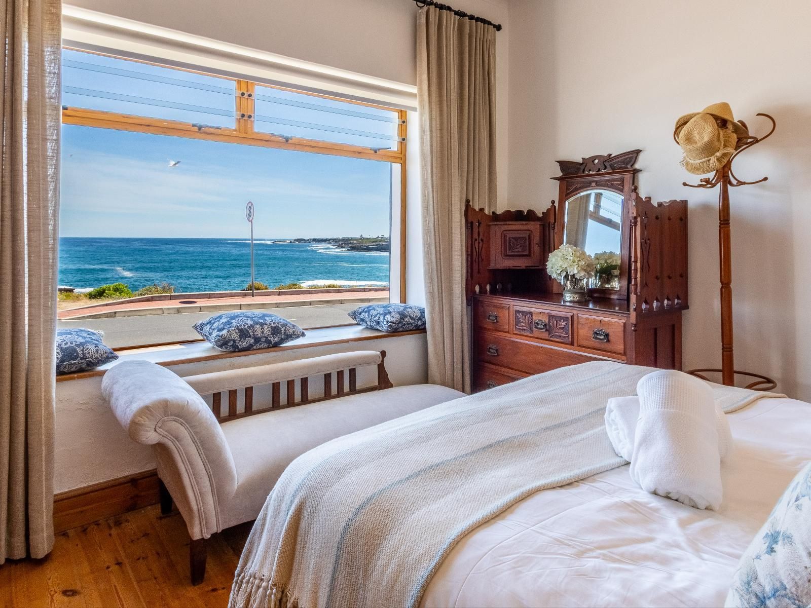 A cozy bedroom featuring a vintage wooden dresser and a large window offering stunning views of the ocean. A comfortable chaise lounge sits by the window, adorned with patterned cushions, enhancing the room's charm and providing a perfect spot for relaxation.
