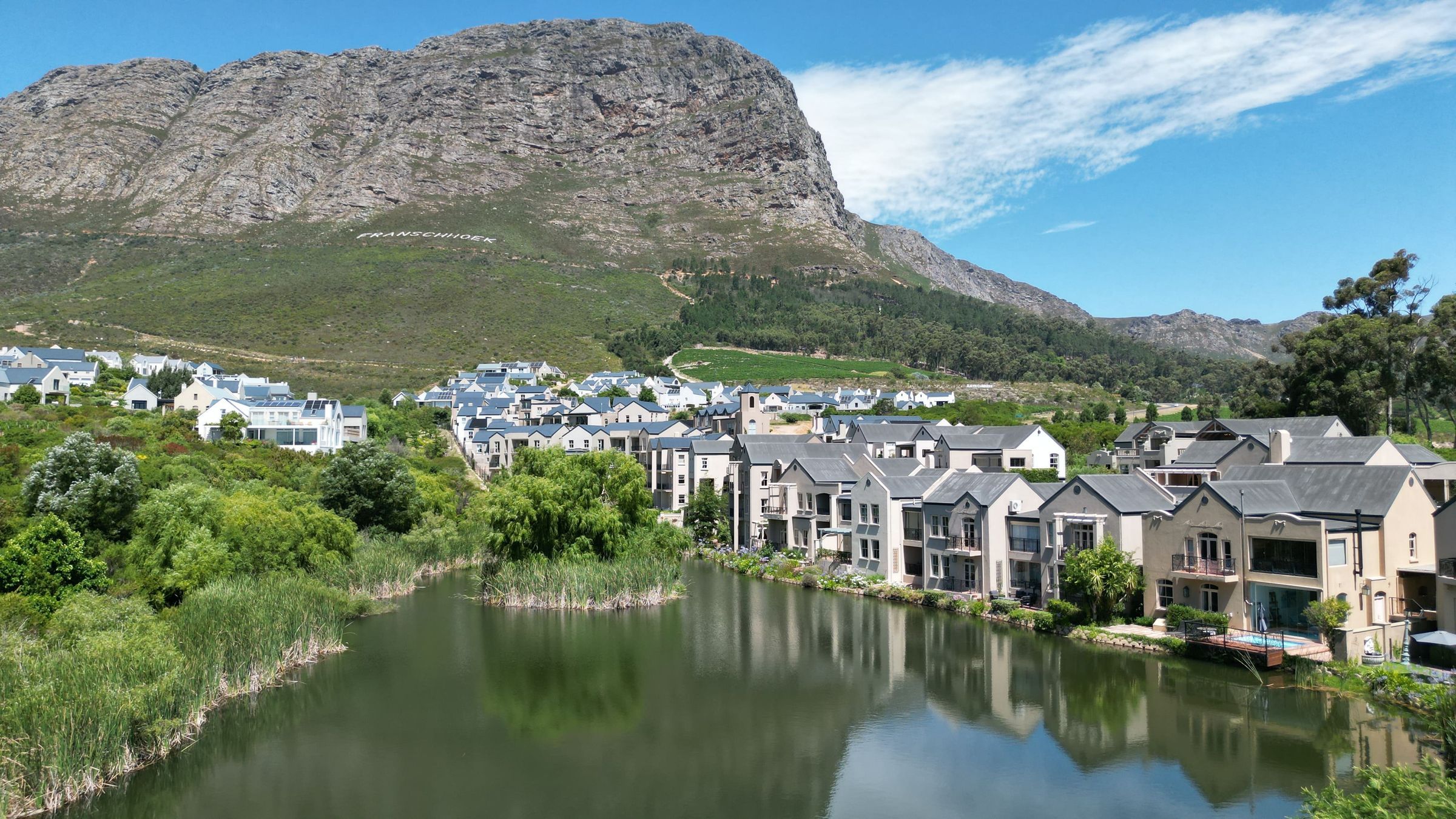 A picturesque view of a charming village nestled by a serene lake, with modern homes reflecting on the water. A lush green landscape surrounds the area, leading up to a rugged mountain backdrop under a clear blue sky.