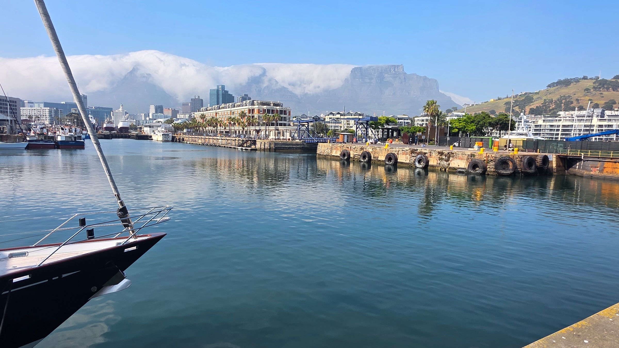 View of V&A Waterfront with a docked sailboat, vibrant shops, and bustling activities against Table Mountain. The scene captures maritime heritage and Cape Town's energy under a clear blue sky.