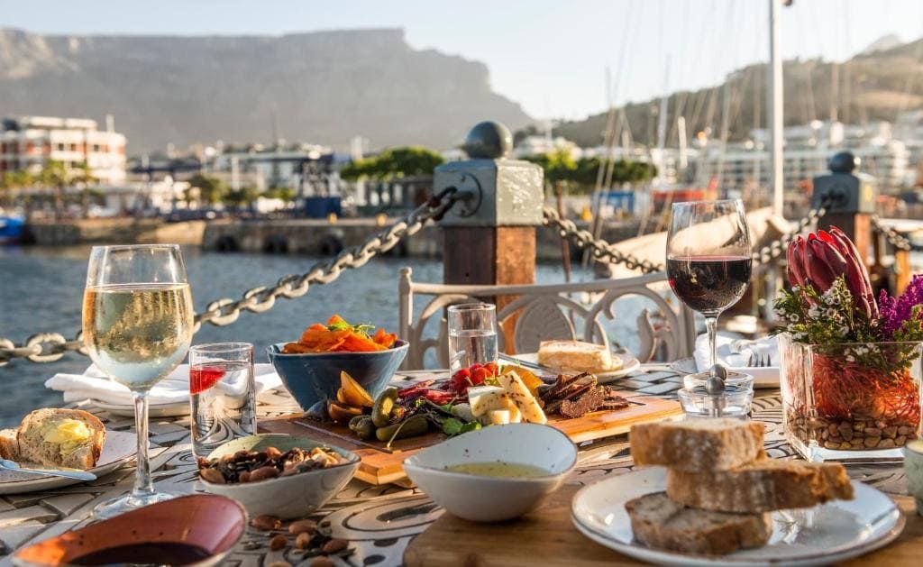 A beautifully set table by the waterfront features an array of dishes, including bread, cheese, and a colorful salad. Two glasses of wine, one red and one white, complement the meal. A floral arrangement adds elegance, with a marina and mountains in the background under a clear sky.