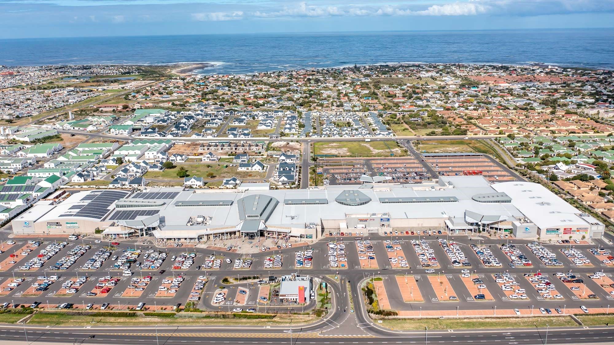 Aerial view of Whale Coast Mall, the central retail destination along the Hermanus coastline. The image highlights the expansive parking area, modern architecture, and its proximity to residential areas and the ocean, showcasing its role as a key shopping location for locals and tourists.