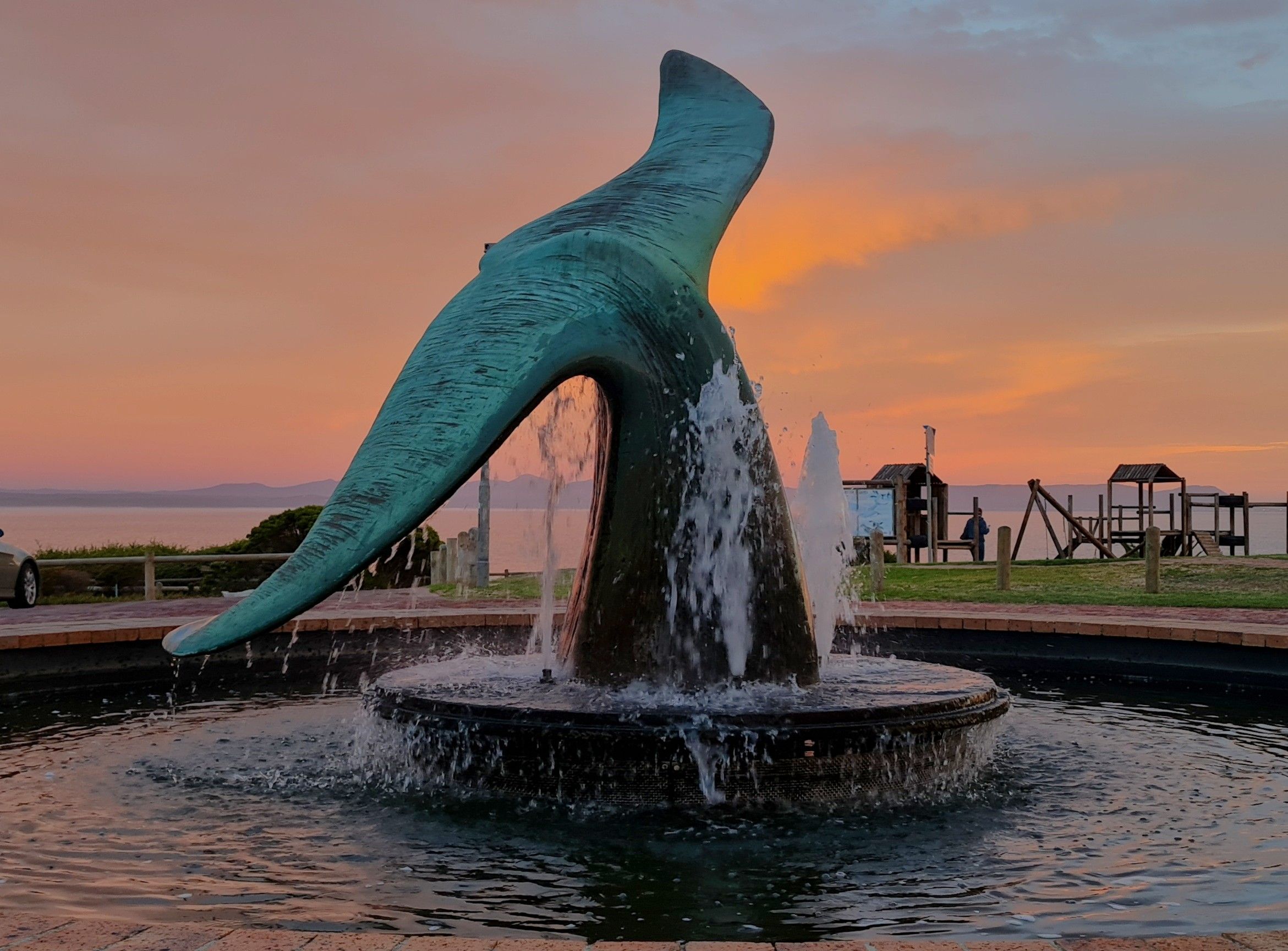 A large, dynamic sculpture of a whale tail emerges from a fountain with water cascading down. The scene is set against a vivid sunset sky, with the ocean and a playground visible in the background, highlighting the artistic blend of nature and creativity in Hermanus.