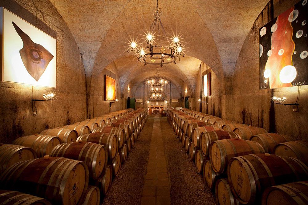 A dimly lit wine cellar with rows of wooden barrels lining both sides. The arched ceiling is adorned with chandeliers, and abstract paintings decorate the stone walls. The cellar has a warm, rustic ambiance and features a distant table set for tasting.