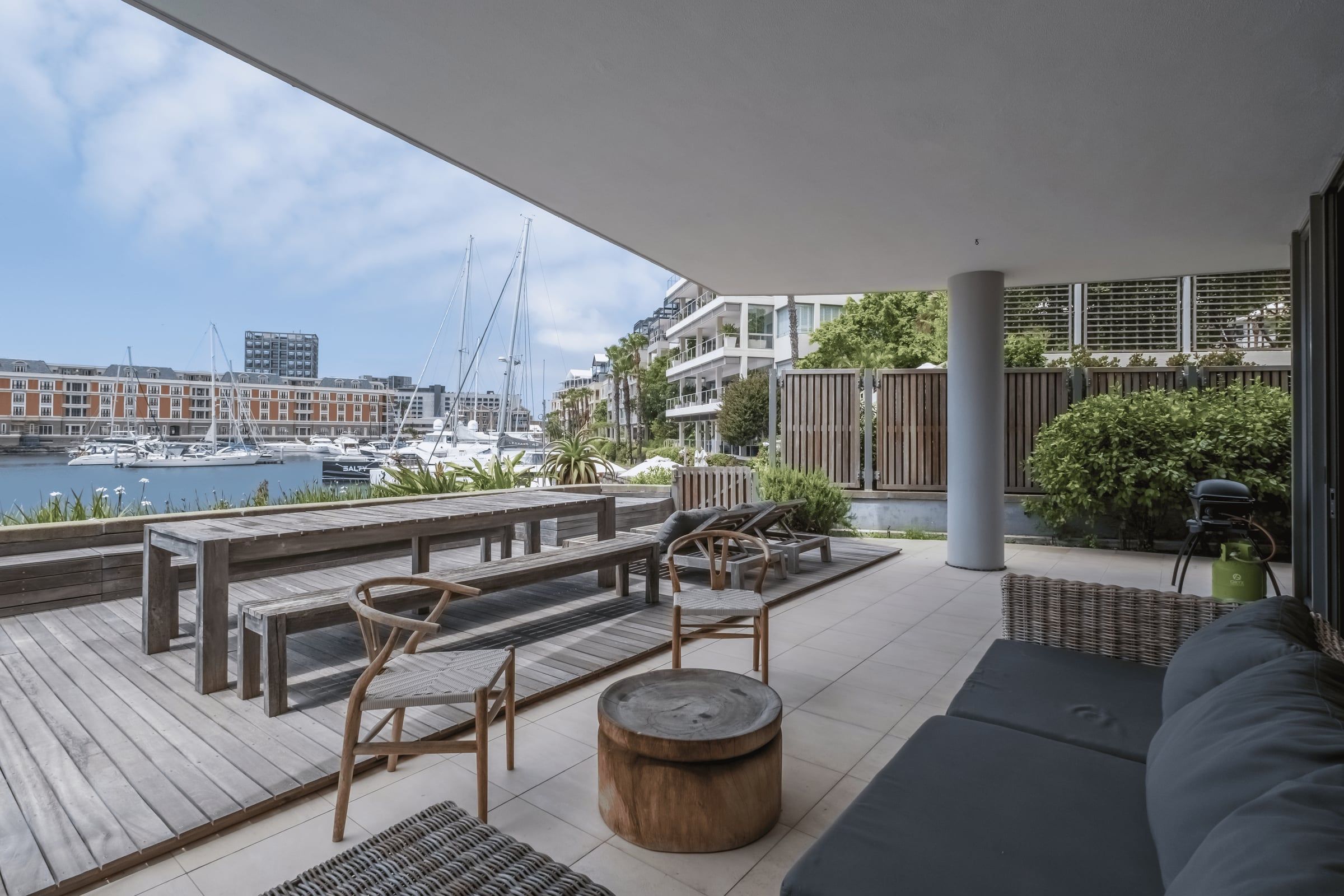 A stylish outdoor patio featuring wicker furniture and wooden benches overlooks a marina with several docked boats. Apartment buildings and lush greenery enhance the serene atmosphere. A grill is visible in the corner, suggesting a space for relaxation and entertaining.